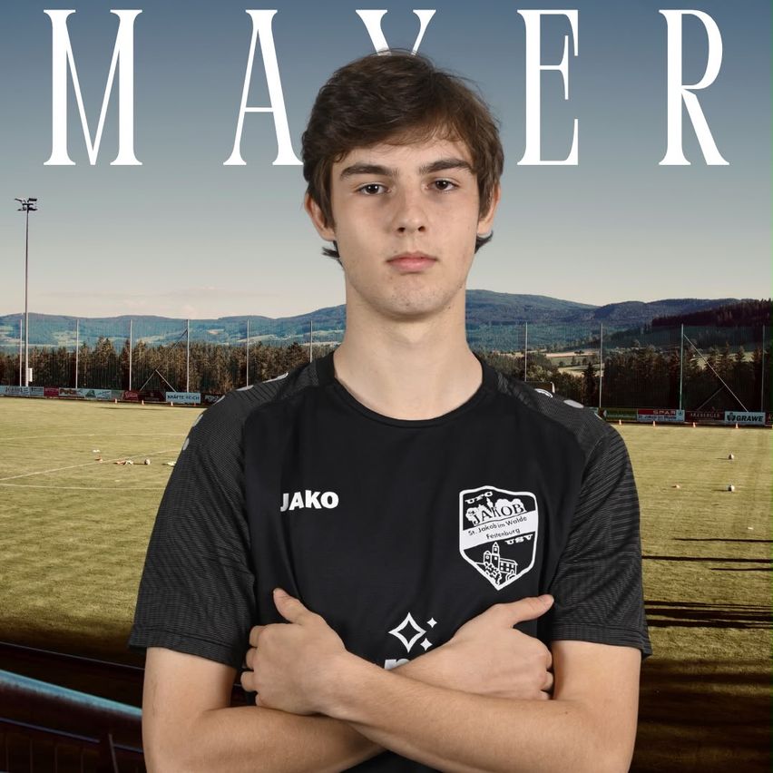 A young man stands confidently with crossed arms in a black soccer jersey with a logo on the left chest. Behind him, a soccer field with a grassy area and mountains in the distance.