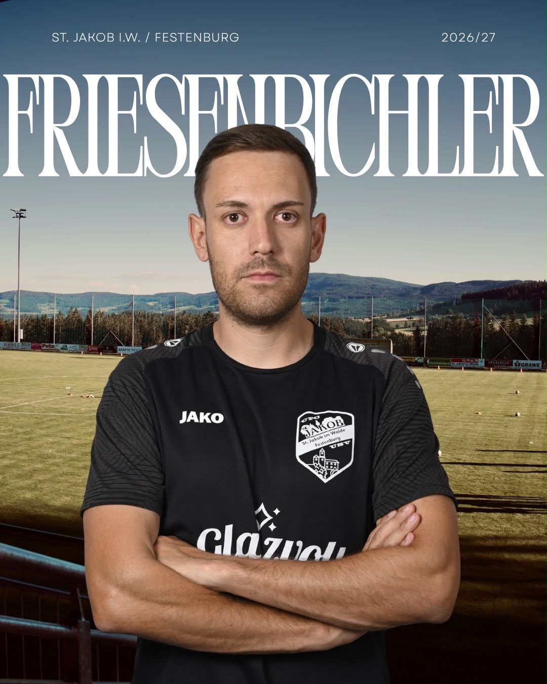 A man stands on a soccer field with his arms crossed, wearing a black jersey with the word 'Jako' and a logo on the left chest. The background features a soccer field with a mountain range and a clear sky.