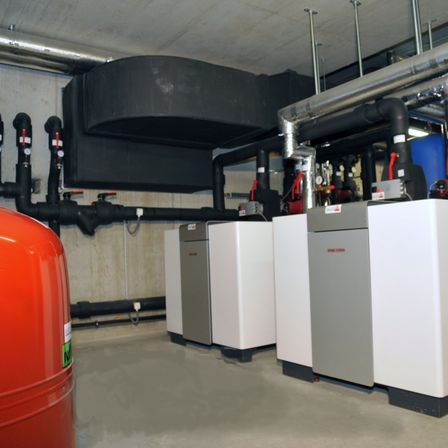 Ein Keller-Nutzraum mit vier weißen und grauen HVAC-Geräten, Rohren und einem großen roten Tank auf der linken Seite.