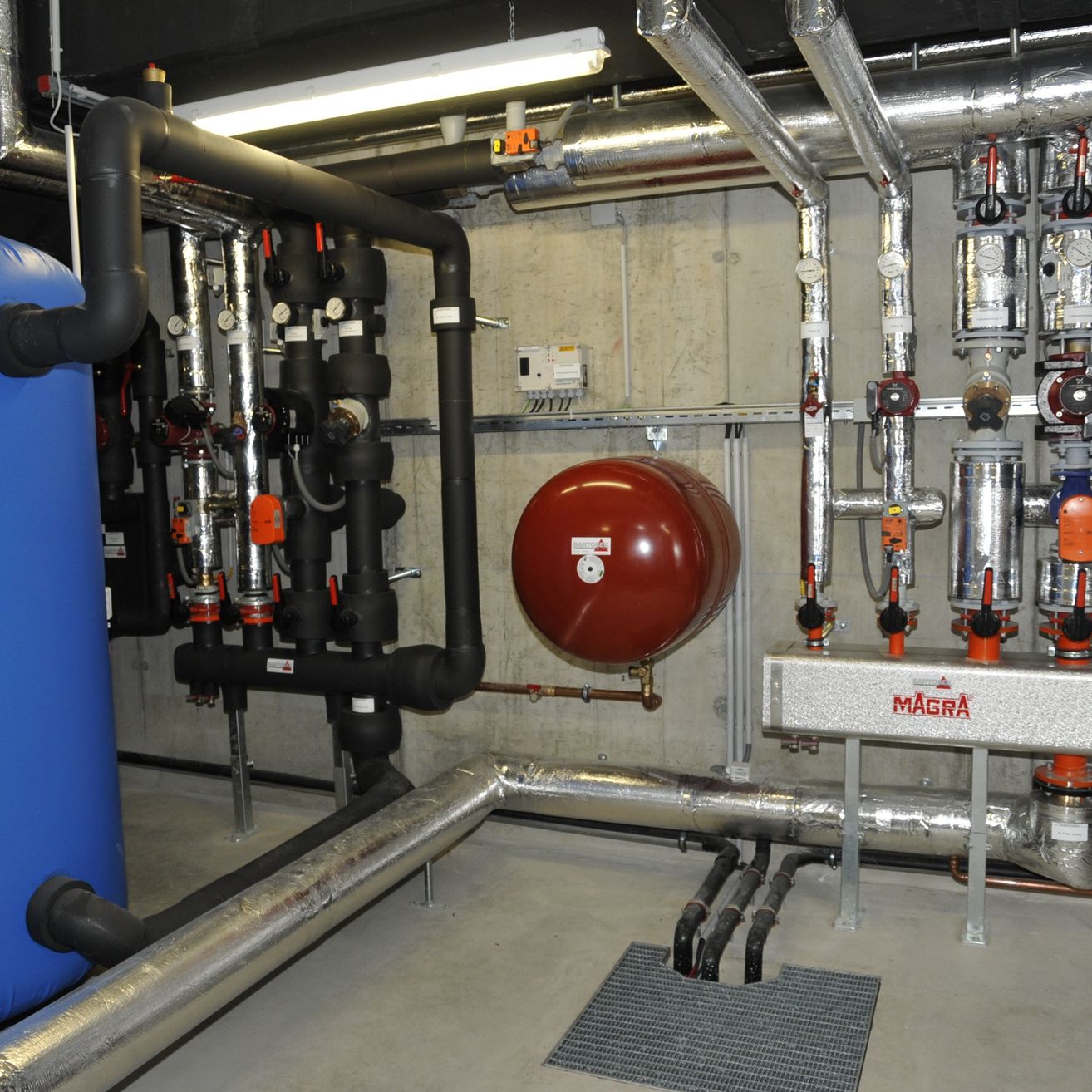 Eine industrielle Anlage mit verschiedenen Rohren und einem zentralen roten Tank, beschriftet mit 'MAGRA', umgeben von zahlreichen Ventilen und Sensoren.