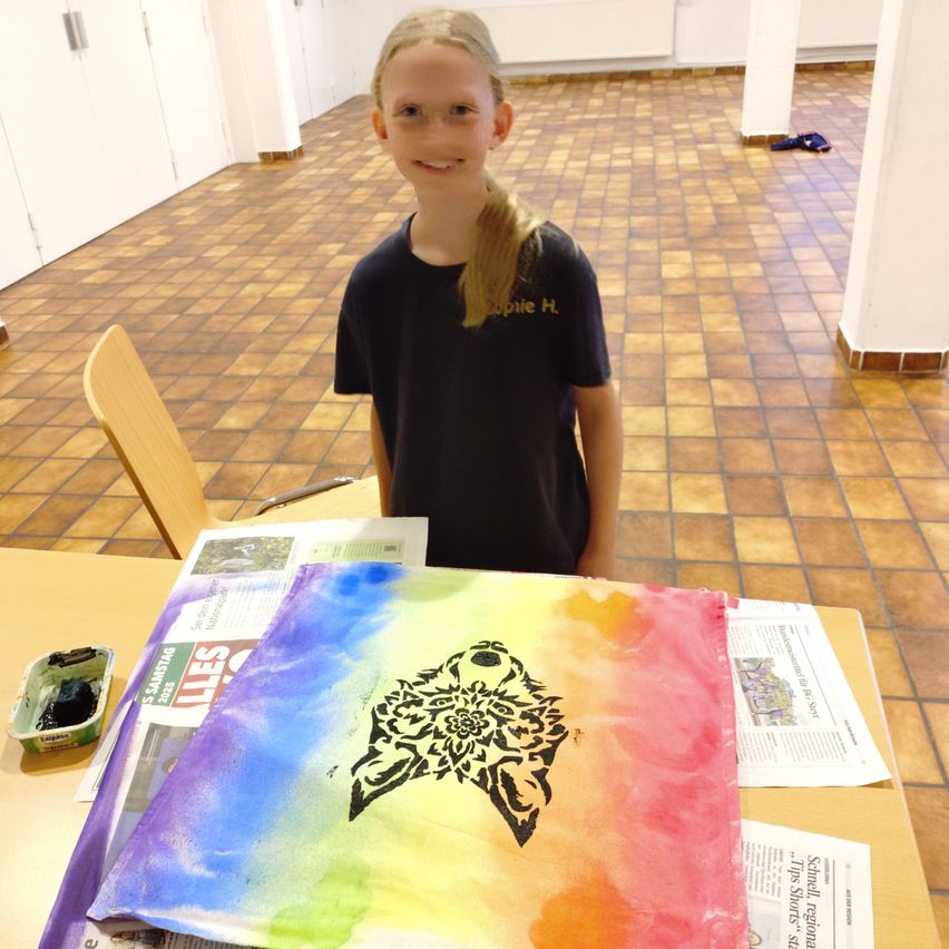 Ein Mädchen steht hinter einem Tisch mit einem farbenfrohen Gemälde darauf. Das Gemälde hat ein schwarzes Design darauf. Sie trägt ein schwarzes T-Shirt mit Text darauf. Hinter ihr ist ein gefliester Boden.