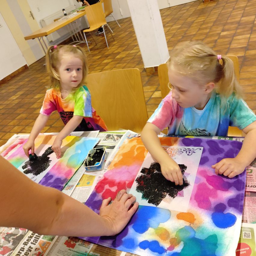 Zwei kleine Mädchen malen auf einer großen Leinwand in einem Raum mit gefliesten Böden. Ein Mädchen benutzt einen Pinsel, während das andere ihre Hände benutzt. Der Tisch ist mit Zeitungen bedeckt.