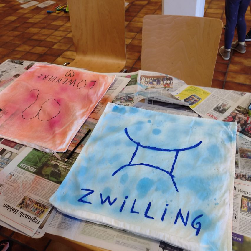 Zwei Stoffstücke sind auf einem mit Zeitungen bedeckten Tisch ausgestellt. Eines zeigt 'TRANSFORMATION' in Schwarz, das andere 'Zwilling' in Blau auf einem blau-weißen Hintergrund. Stühle und eine Person sind im Hintergrund zu sehen.