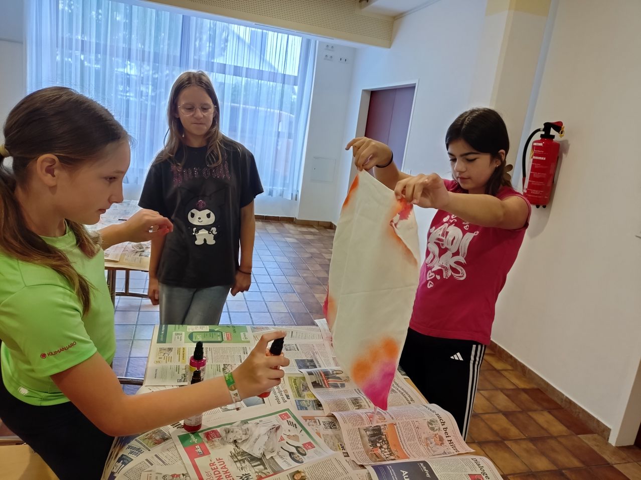 Drei Mädchen arbeiten an einem Batik-Projekt an einem Tisch. Eine hält ein weißes Tuch, eine andere eine Flasche mit Farbe. Ein Feuerlöscher ist an der Wand hinter ihnen angebracht.
