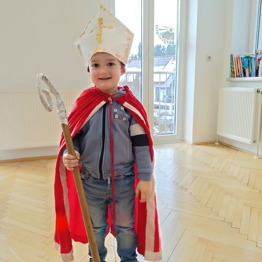 Ein junger Junge in einem roten Umhang und einer weißen Mütze mit goldenen Verzierungen, die einen Stab hält, steht auf einem Holzboden in einem Raum.