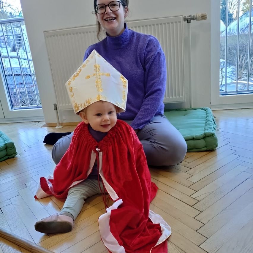 Eine lächelnde Frau in einem lila Pullover sitzt mit einem Kind, das einen roten Umhang und einen golden-weißen Bischofshut trägt, auf dem Boden.