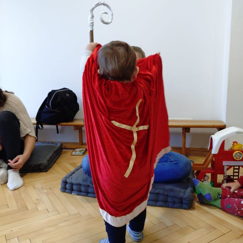 Ein Kind in einem roten Gewand mit einem Kreuz steht in einem Raum und hält einen Stab. Hinter ihm sitzt eine Frau auf dem Boden. In der Nähe sind ein Rucksack und Spielzeug zu sehen.