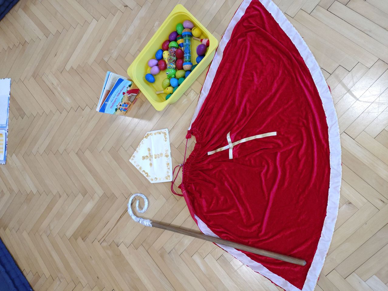 Ein roter Samtrock, ein Buch, eine gelbe Spielzeugkiste mit bunten Eiern, ein weißer Stoff mit Kreuzdruck und ein Holzstab liegen auf einem Holzboden aus.