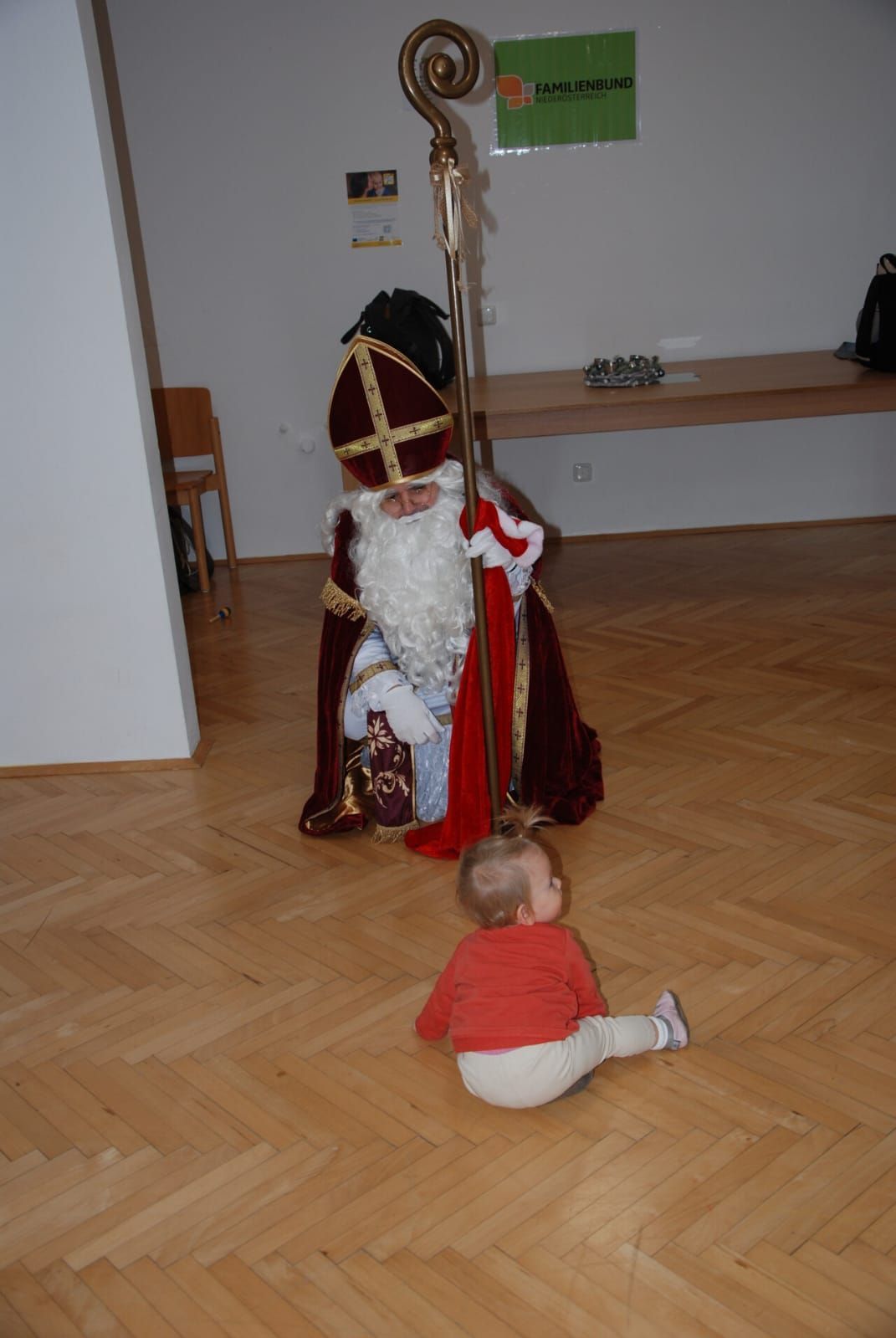 Ein Mann in der Kleidung des Heiligen Nikolaus kniet neben einem kleinen Kind in einem roten Shirt auf einem Holzboden.