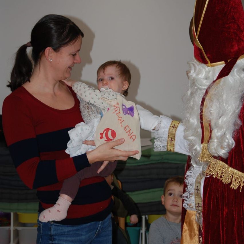Eine Frau hält ein Baby mit Schnuller und einem Tuch mit der Aufschrift 'Natal Ena'. Eine Figur von Santa Claus überreicht dem Baby ein Geschenk. Ein weiteres Kind steht in der Nähe.