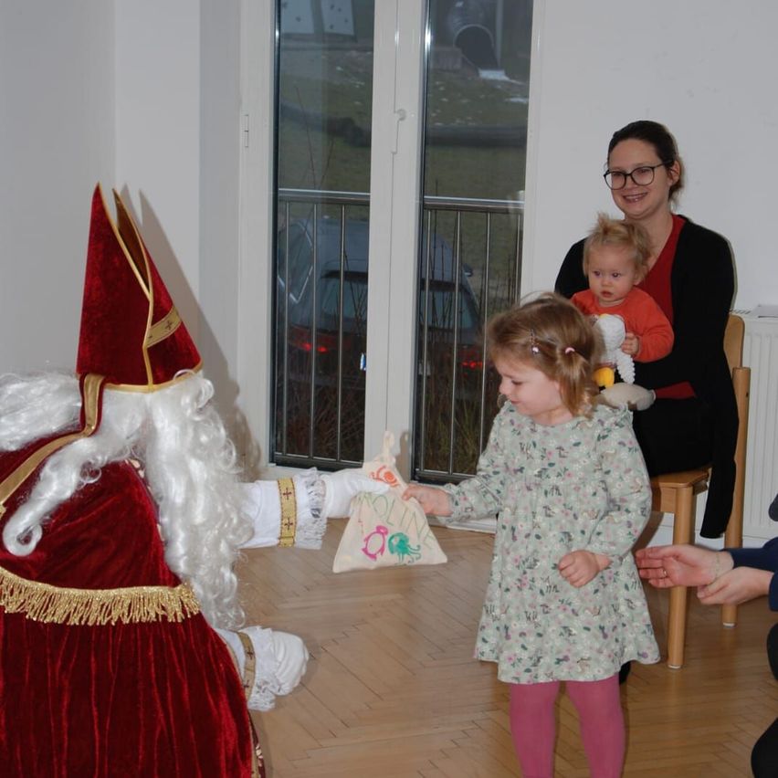 Eine als Weihnachtsmann verkleidete Person gibt einem kleinen Mädchen ein Geschenk, während eine Frau ein Baby hält und eine andere Person deren Hand hält in einem Raum mit einer Glastür.