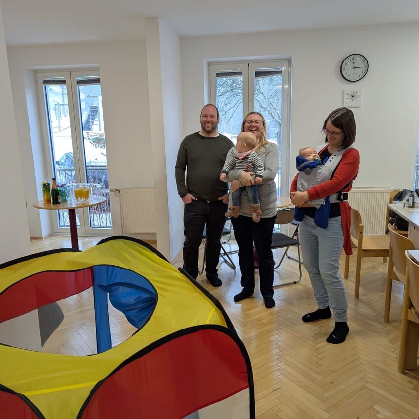 Vier Erwachsene stehen in einem Raum mit jeweils zwei Babys. Ein farbenfrohes Zelt steht im Vordergrund. Dahinter steht ein Tisch mit Getränken und Stühle um einen Tisch herum. Fenster mit Blick auf verschneite Bäume sind auf der linken Seite.