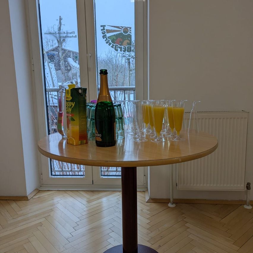 Ein holzner Tisch hat eine Flasche Sekt, Orangensaft und Champagner-Gläser. Dahinter führt eine Glastür mit einem grünen Logo zu einem verschneiten Ausblick.