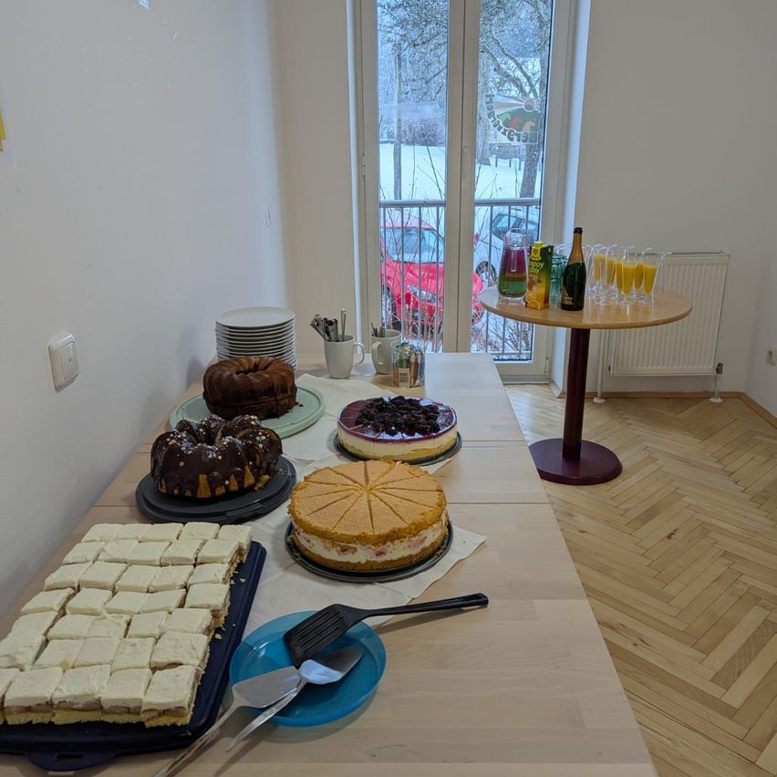 Ein Tisch voller verschiedener Desserts, darunter ein Gugelhupf, Käsekuchen und andere Gebäckstücke, vor dem Hintergrund eines Zimmers mit Balkon und verschneiter Aussicht.