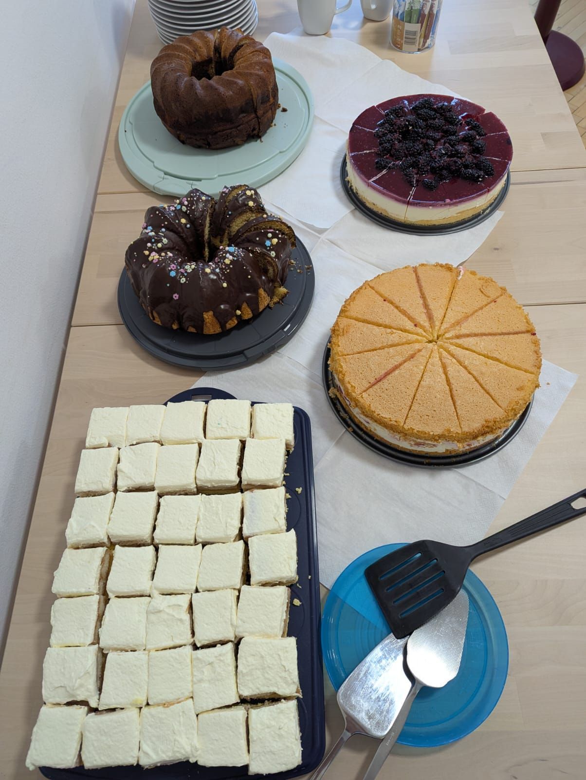 Ein Holztisch zeigt verschiedene Desserts. Eines ist ein Schokoladen-Bundt-Kuchen, ein anderes ist ein Käsekuchen mit Brombeeren. Ebenfalls vorhanden sind ein Stück Käsekuchen und eine Platte mit quadratischen Desserts.