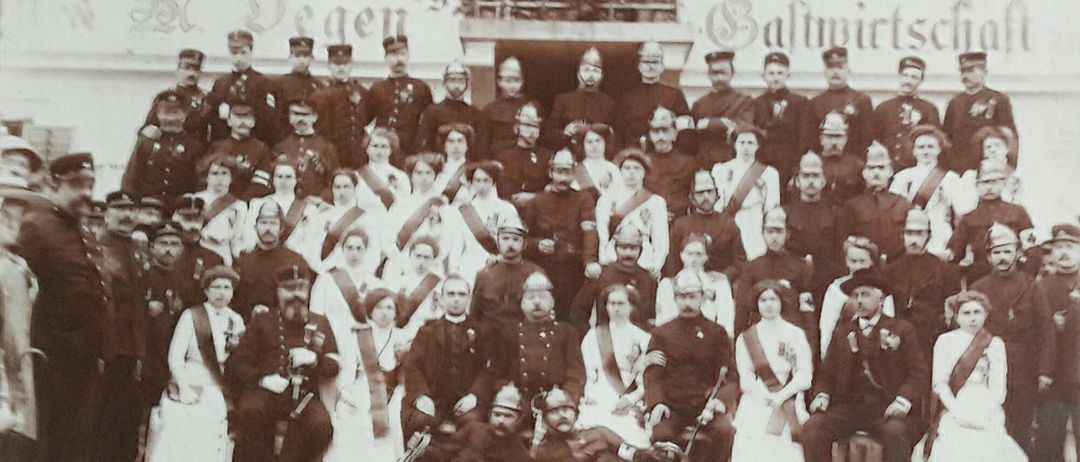 Eine Gruppenaufnahme von uniformierten Personen, einige sitzend und einige stehend, vor einem Gebäude mit der Aufschrift 'Salzwirts'. Ein Mann im Vordergrund liegt auf dem Boden und hält ein Gewehr.