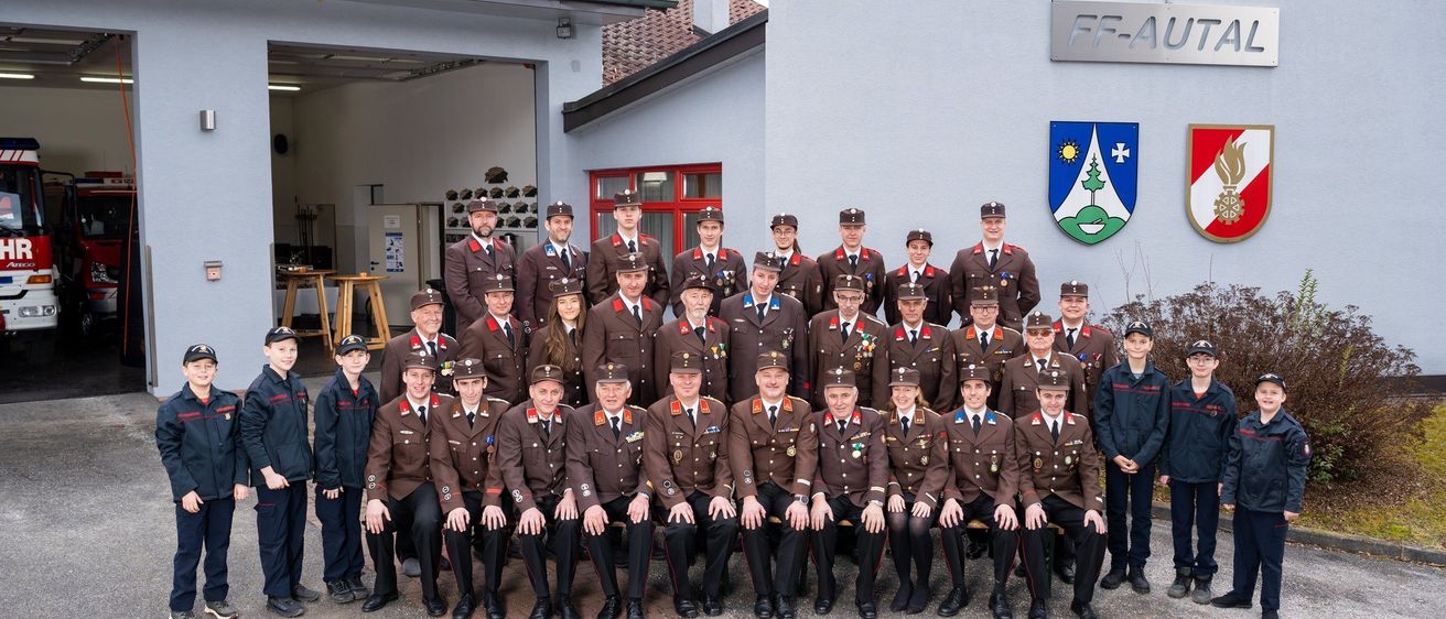 Eine Gruppe uniformierter Personen steht vor einem Gebäude mit einem Emblem und einem Schild. Die Uniformen haben Abzeichen und Medaillen. Sie lächeln für ein Foto.