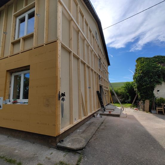 Ein Außenansicht eines Hauses im Bau mit Holzwänden und gelber Isolierung. Es gibt Fenster, eine Leiter und Werkzeuge in der Nähe des Hauses.