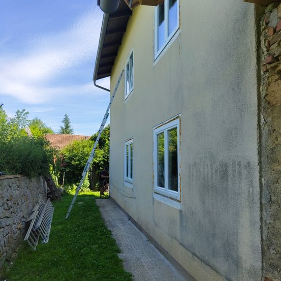 Ein Haus mit gelben Wänden und drei Fenstern. Eine Leiter lehnt an das Haus. Eine Person steht in der Nähe.