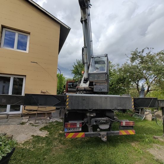 Ein Kran steht vor einem beigen Haus mit Glasfenstern unter einem bewölkten Himmel.