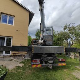 Ein Kran steht vor einem beigen Haus mit Glasfenstern unter einem bewölkten Himmel.