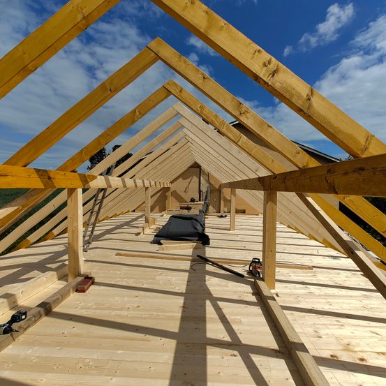 Ein hölzerner Dachbodenraum im Bau. Holzbalken und eine Leiter sind sichtbar. Ein schwarzes Objekt liegt auf dem Boden.