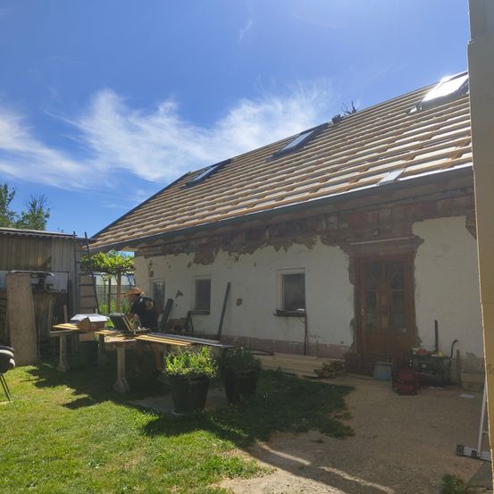 Ein Haus im Bau mit einem neuen Dach und einem Mann, der draußen an einem Tisch arbeitet. Werkzeuge und Materialien sind herum verstreut.