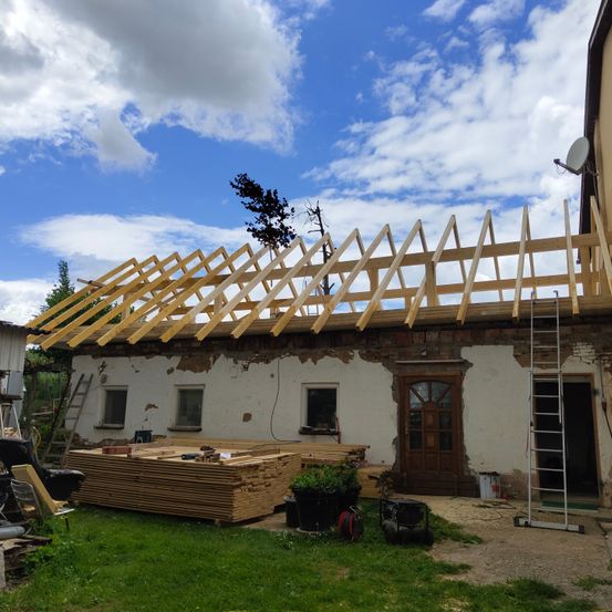 Ein Haus im Bau mit einem hölzernen Dachgerüst, mehreren Leitern, Holzbrettern, einer Satellitenschüssel und einer Tür. Das Haus hat eine Ziegelwand mit etwas abblätternder Farbe.