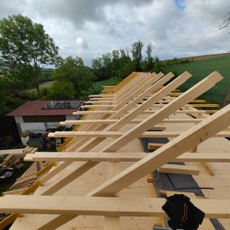Ein Blick von oben auf ein Gebäude im Bau mit Holzbalken, die die Dachkonstruktion bilden. Solarpanele sind auf dem Dach installiert. Bäume und ein grünes Feld sind im Hintergrund zu sehen.