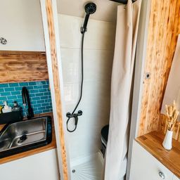 Kleines Badezimmer mit weißen Fliesen und einer Dusche. Ein silbernes Waschbecken mit schwarzem Wasserhahn steht neben der Dusche. Ein weißer Vase mit Trockenblumen steht auf dem Counter.