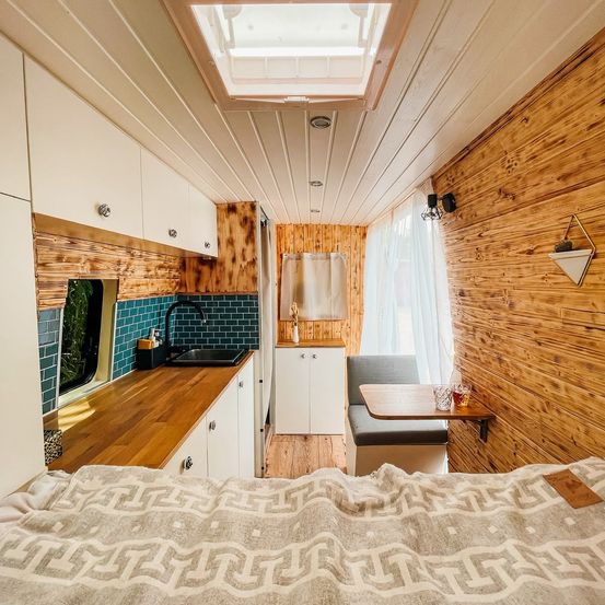 Innenraum eines Wohnmobils mit Holzwänden und einer kleinen Küchenzeile. Im Vordergrund ist ein Bett zu sehen.