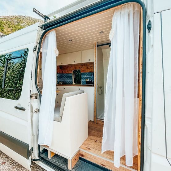 Der Innenraum eines weißen Wohnmobils mit geöffneter Tür. Im Inneren befindet sich ein kleiner Küchenbereich mit gefliesten Wänden, einer Dusche und einer Bankähnlichen Sitzgelegenheit.