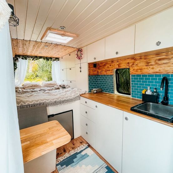 Ein gemütliches Interieur eines Wohnmobils mit Bett, Küche und Bad. Das Bett hat Kissen und eine Decke. Die Küche hat ein Waschbecken, einen Wasserhahn und ein kleines Fenster. Das Badezimmer hat eine Dusche.