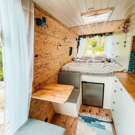 Innenraum eines Wohnmobils mit Holzwänden, einem Bett, einem Tisch und einem Fenster. Das Bett hat Kissen, eine Matratze und einen kleinen Teppich.
