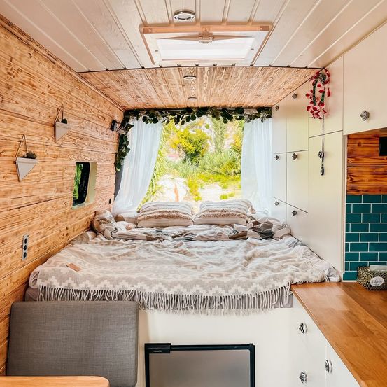 Ein gemütliches Wohnmobil-Interieur mit Holzwänden, einem Bett und einer Küchenzeile. Das Bett ist mit einer Decke bedeckt, und es gibt Kissen und ein Fenster mit weißen Vorhängen.