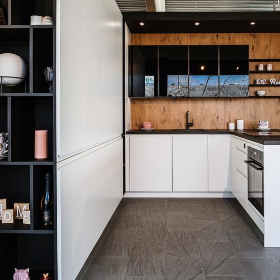 Eine moderne Küche mit weißen Schränken und Holzwänden, ausgestattet mit einer schwarzen Arbeitsplatte, einem Waschbecken mit Wasserhahn und einem eingebauten Ofen. Verschiedene Gegenstände sind auf Regalen und Schränken platziert.