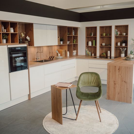 Moderne Küche mit weißen Schränken, Holzregalen und einem grünen Stuhl auf einem runden Teppich. Ein Holztisch hält ein Buch, während die Arbeitsplatte verschiedene Küchenutensilien zeigt.