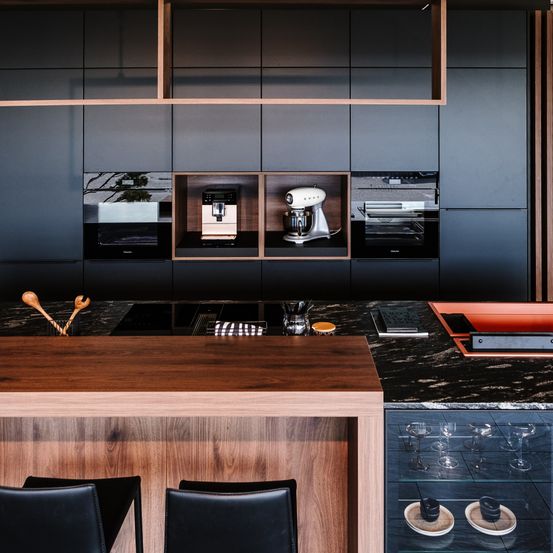 Eine moderne Küche mit schwarzen Schränken, einer hölzernen Arbeitsplatte und verschiedenen Küchengeräten. Sie hat schwarze Marmorarbeitsplatten, zwei Stühle und einen Weinglasschrank.