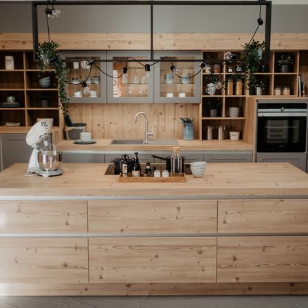 Eine moderne Küche mit Holzschränken und Arbeitsplatten. Ein Mixer und eine Ablage mit Flaschen und Gläsern stehen auf der Insel. Regale mit verschiedenen Gegenständen sind an der Wand angebracht.