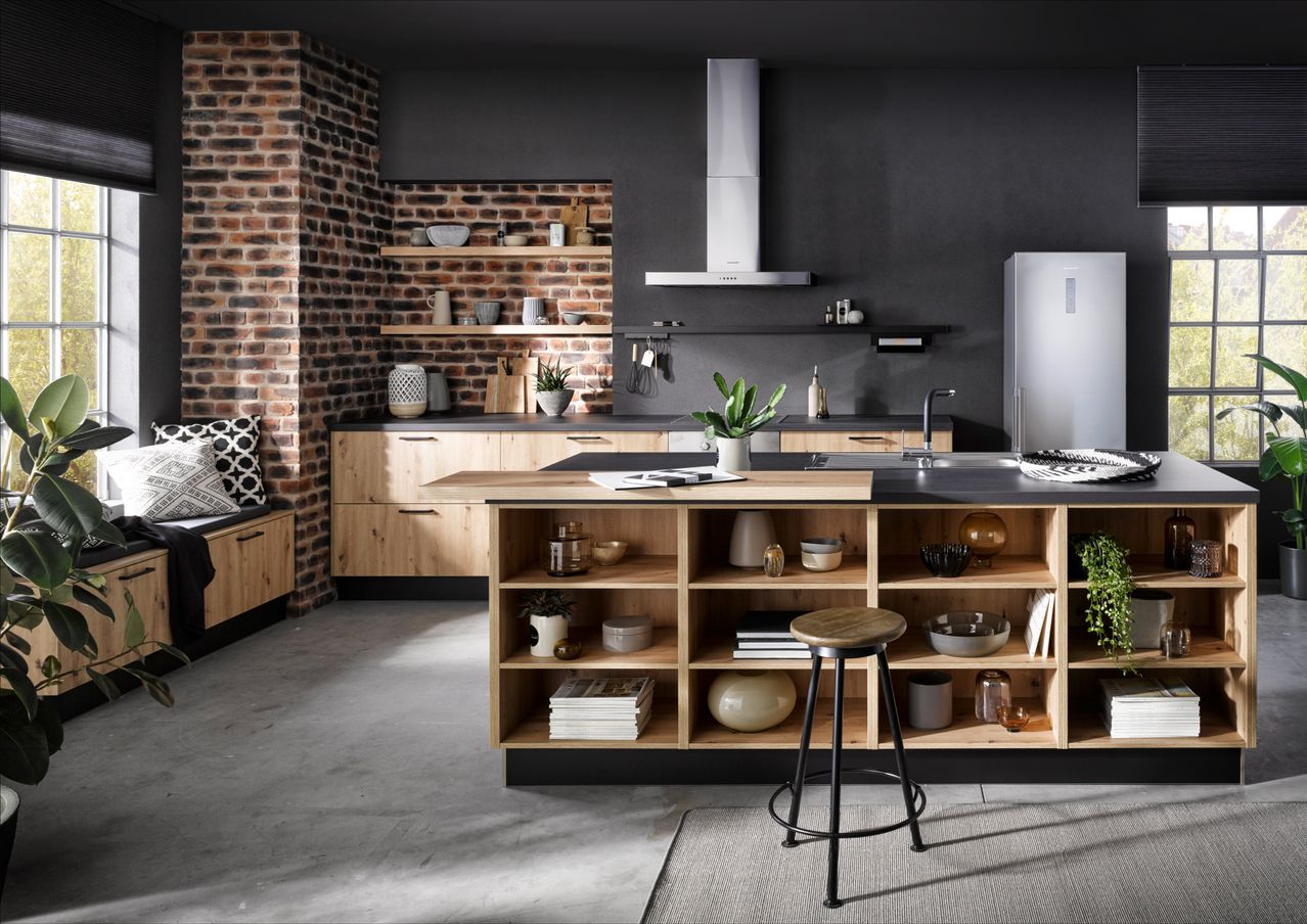 Eine moderne Küche mit Holzschränken und einer schwarzen Ziegelwand. Ein hölzerner Tresen mit einem Hocker, einem Kühlschrank und einem Waschbecken mit Wasserhahn. Regale mit verschiedenen Gegenständen wie Büchern, Pflanzen und Dekorationsartikeln.