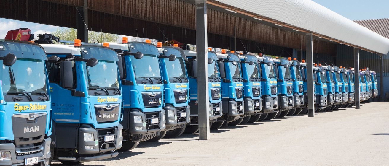 Eine Reihe blauer MAN-Lkw ist unter einer großen Metallstruktur mit gewölbtem Dach geparkt. Die Lkw sind mit orangefarbenen Lichtern ausgestattet. Der Himmel ist klar mit einigen Wolken.