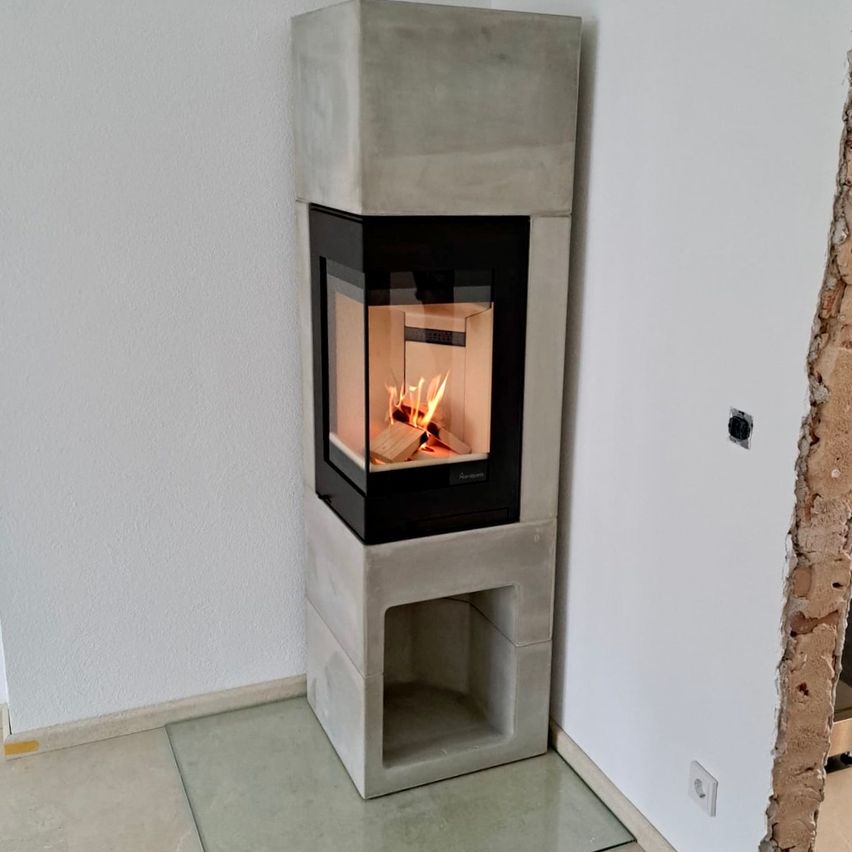 Ein moderner Betonkamin mit brennendem Feuer steht in einer Ecke eines Raumes mit weißer Wand. Der Kamin hat ein Glasfenster.
