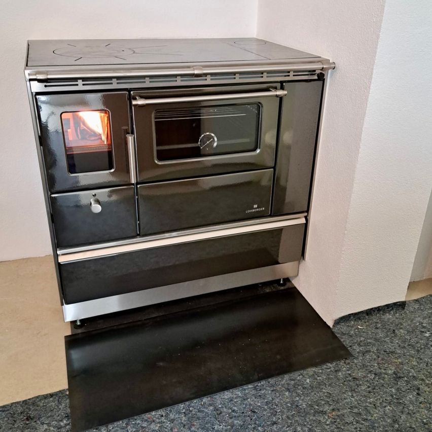 Ein moderner, schlanker, edelstahlfarbener Doppelofen mit brennendem Feuer im Ofen und einer Glastür, der auf einem schwarzen Teppich in einer Ecke eines Raumes steht.