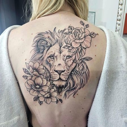 Ein detailliertes Tattoo eines Löwenkopfes mit Blumen auf dem Rücken einer Frau. Der Löwe hat eine Feder in seiner Mähne.