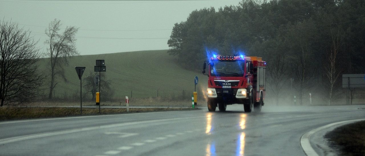Ein roter Feuerwehrwagen mit blauen Lichtern fährt auf einer nassen Straße. Im Hintergrund befinden sich Bäume und Stromleitungen.