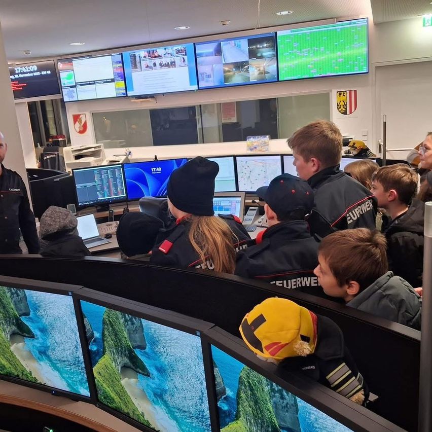 Eine Gruppe von Kindern und Erwachsenen versammelt sich um einen Computerarbeitsplatz, möglicherweise in einem Kontrollraum. Sie betrachten Monitore, die verschiedene Bilder und Informationen anzeigen. Die Kinder tragen Mützen und die Erwachsenen Uniformen.