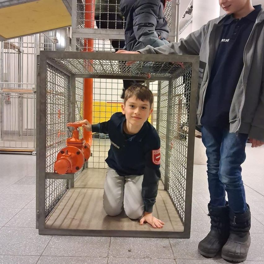 Ein junger Junge kniet in einem Metallkäfig, ein anderer Junge hält sich am Käfig fest. Sie tragen beide Winterkleidung. Hinter ihnen steht eine Person in der Nähe eines Käfigs.