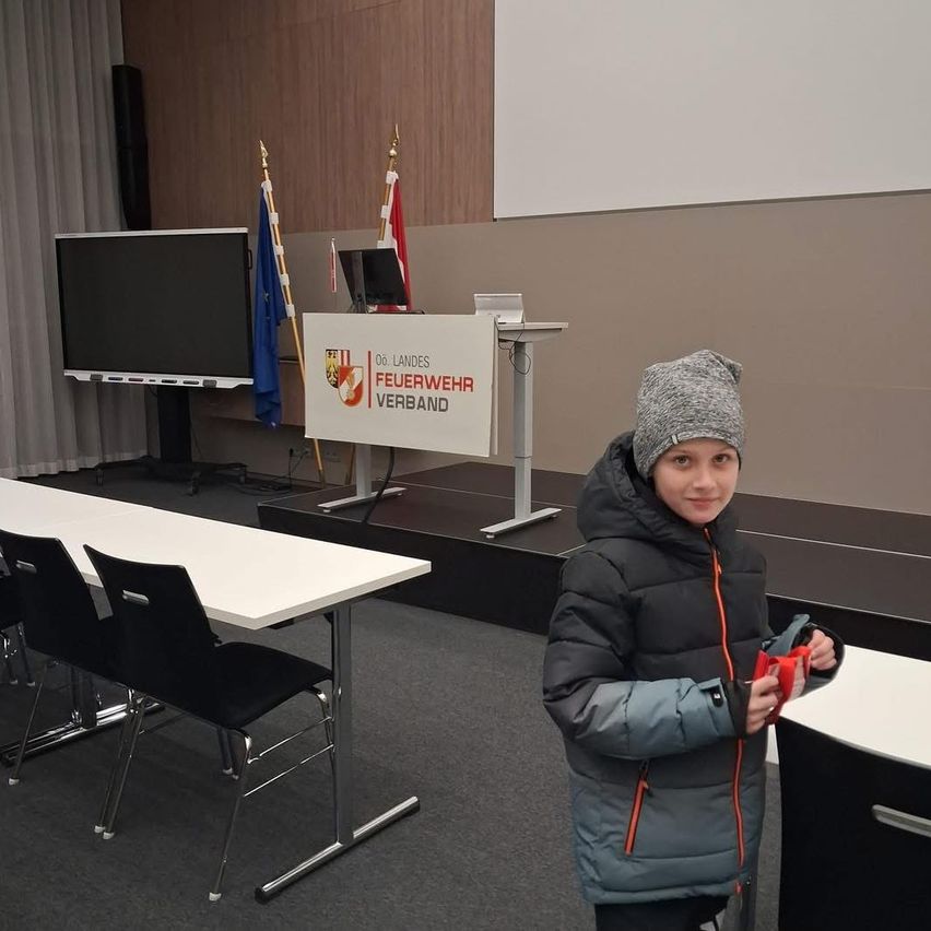 Ein Kind steht in einem Konferenzraum und hält einen roten Gegenstand in der Hand, hinter ihm steht ein Podium mit Flaggen, links ein Bildschirm und rechts eine Tafel.