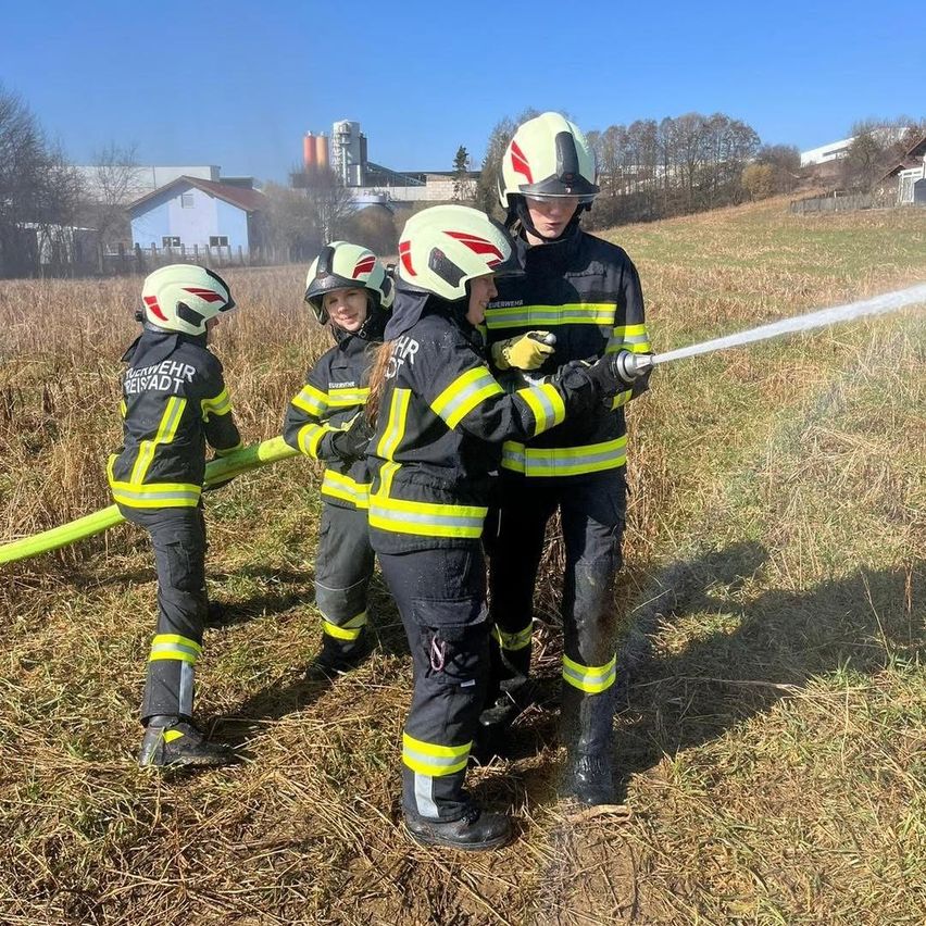 Vier junge Feuerwehrleute in Uniformen üben das Wassersprühen auf einem Feld. Bäume und Gebäude sind im Hintergrund.
