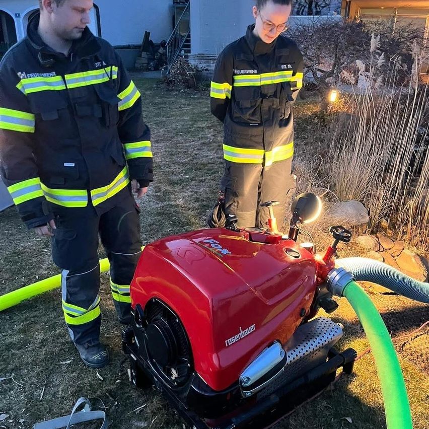 Zwei Feuerwehrleute in Schutzkleidung stehen neben einem roten Rosenbauer-Fahrzeug, das mit einem Schlauch verbunden ist. Sie befinden sich in einem Garten mit einem Haus und Treppen im Hintergrund.
